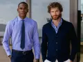 Dos hombres posan. Uno lleva una camisa de vestir azul con corbata, la camisa por dentro. El otro, una camisa de rayas azules y blancas con cárdigan azul oscuro, la camisa por fuera.