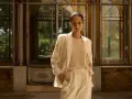Mujer con blazer beige y pantalones de vestir, de pie frente a una estructura con ventanas.