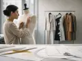 Modista ajusta un vestido en un maniquí, rodeada de patrones y prendas de **marcas pret a porter** colgadas en perchas.