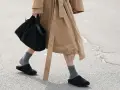 Persona camina con zuecos negros y calcetines grises, llevando un bolso negro y un abrigo beige.