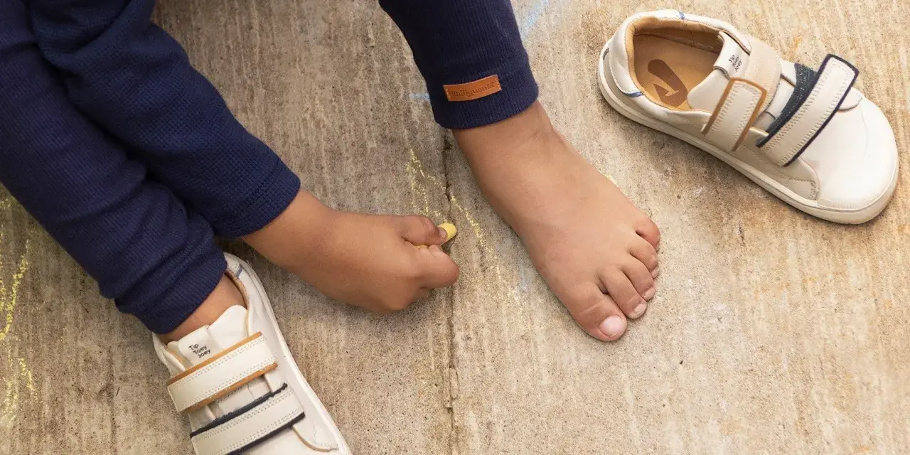 Niño descalzo dibuja con tiza junto a sus zapatos de calzado fisiológico, listos para la aventura.