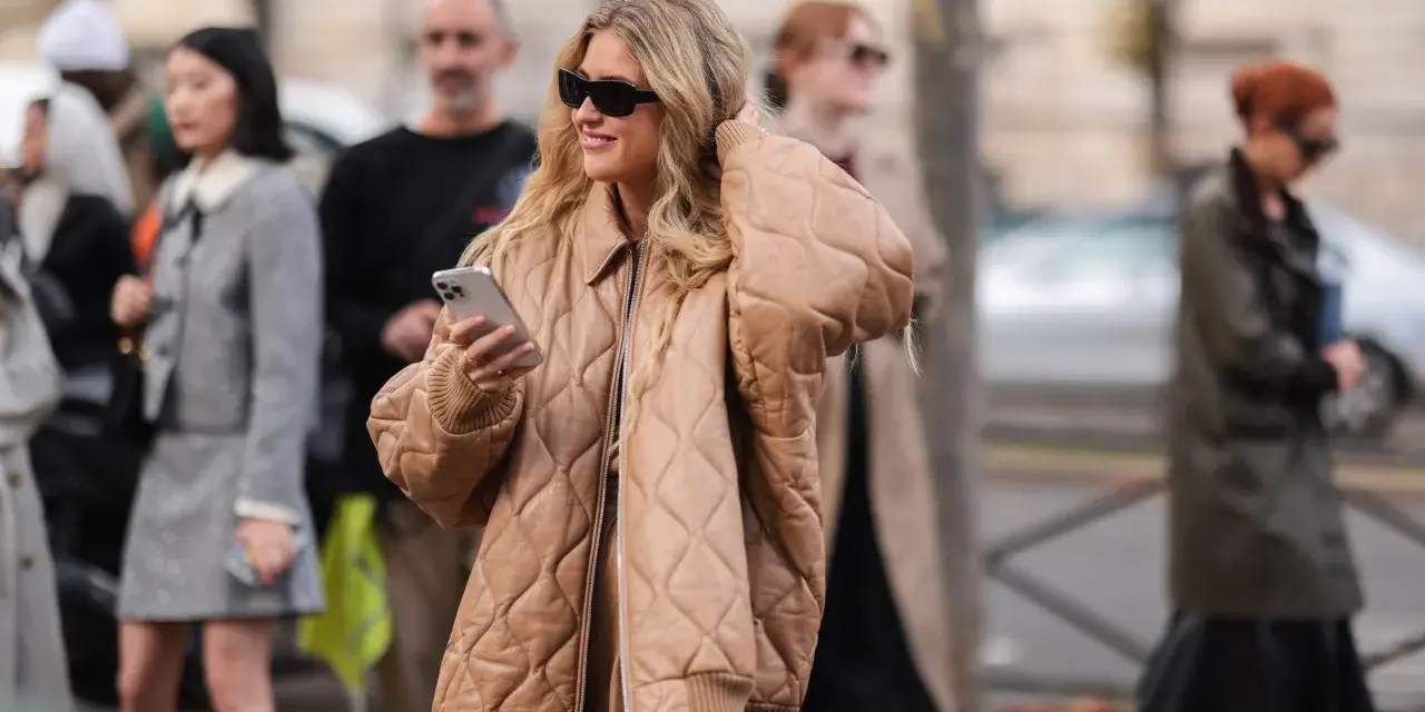 Mujer sonriente con gafas de sol y un abrigo beige claro acolchado, sosteniendo un teléfono.