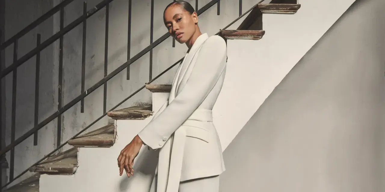 Mujer posando con un elegante outfit blazer blanca, pantalón a juego y cinturón, junto a una escalera antigua.