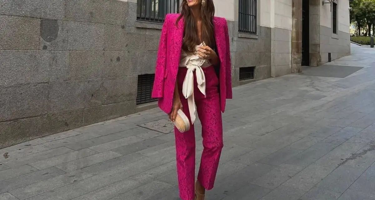 Mujer sonriente luce un espectacular outfit rosa de traje pantalón y blazer, con un cinturón blanco y bolso a juego.
