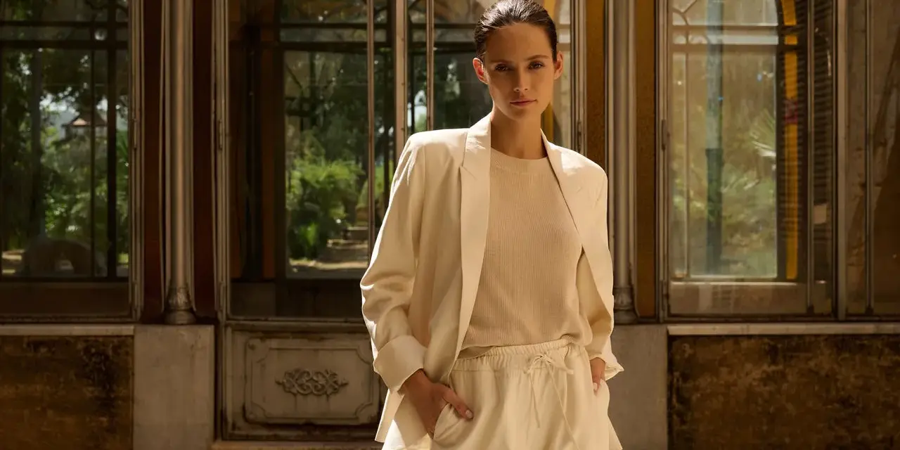 Mujer con blazer beige y pantalones de vestir, de pie frente a una estructura con ventanas.