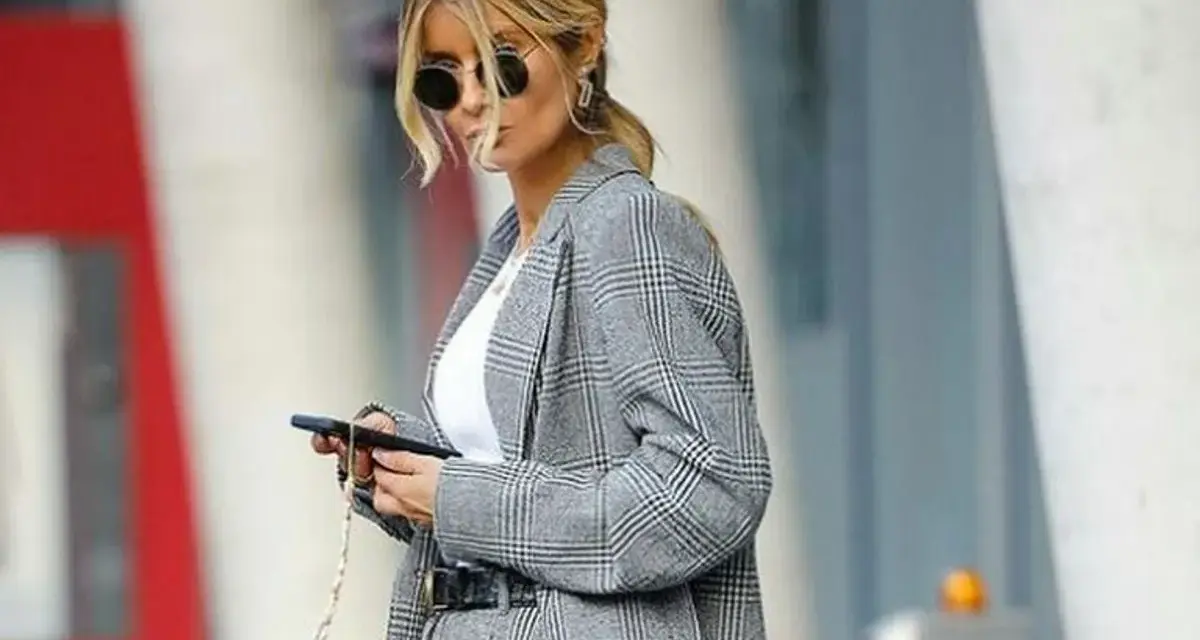 Una mujer con gafas de sol y un traje de cuadros grises, con el pelo recogido, parece una profesora secundaria revisando su móvil.