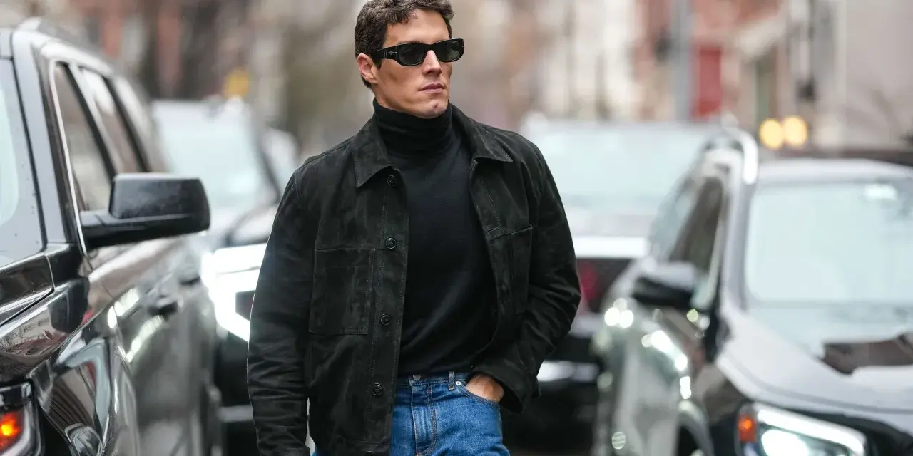 Hombre con gafas de sol y chaqueta de gamuza negra sobre jersey de cuello alto, demostrando como tener estilo.