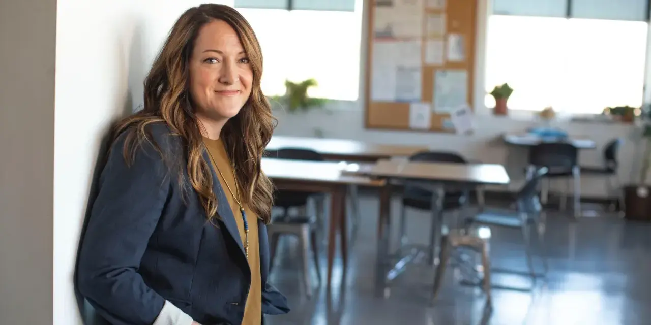 Profesora joven sonriente, apoyada en una pared blanca, con un blazer azul marino y un collar llamativo. El aula al fondo está lista para la clase.