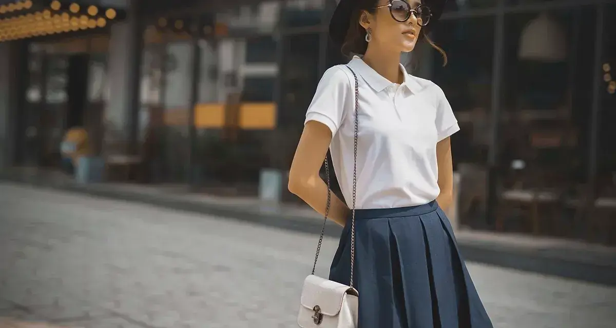 Mujer con estilo preppy, luce un polo blanco, falda azul y sombrero negro.