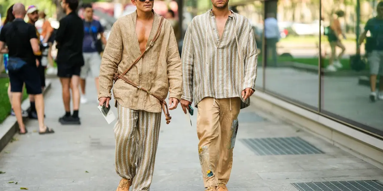 Dos hombres pasean con estilo. Uno lleva un kimono beige y pantalones de lino a rayas. El otro, una camisa a rayas y pantalones beige. Un ejemplo de cómo combinar pantalón lino beige hombre.