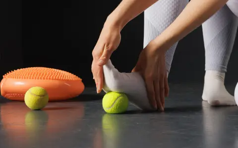 Manos masajeando pie con pelota de tenis, parte de ejercicios fascitis plantar. A su lado, un disco de equilibrio naranja.