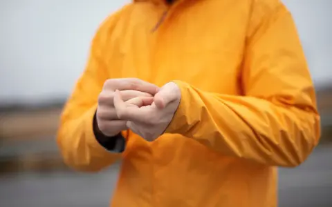 Persona se frota las manos dentro de una chaqueta naranja brillante. El poliéster da calor y protege del frío.