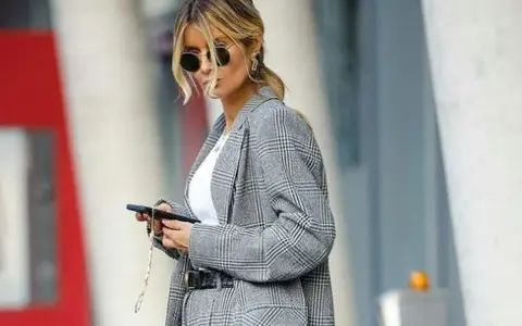 Una mujer con gafas de sol y un traje de cuadros grises, con el pelo recogido, parece una profesora secundaria revisando su móvil.