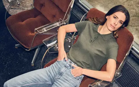 Mujer con vaqueros y polo verde, sentada en una silla de terciopelo, encarnando la moda atemporal.