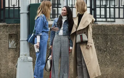 Tres mujeres conversan, una con un traje completo de **denim**, otra con chaqueta **denim** y pantalones a cuadros, y la tercera con un abrigo beige y pantalones a cuadros.