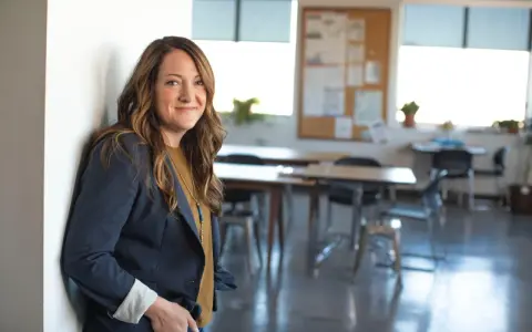 Profesora joven sonriente, apoyada en una pared blanca, con un blazer azul marino y un collar llamativo. El aula al fondo está lista para la clase.