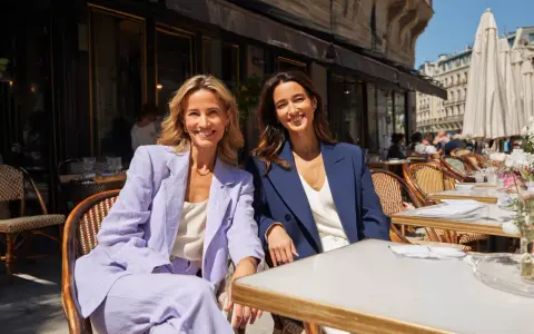 Dos mujeres sonríen sentadas en un café. Una lleva un traje lila, la otra uno azul marino. Ideas para como vestirse para una comunión.