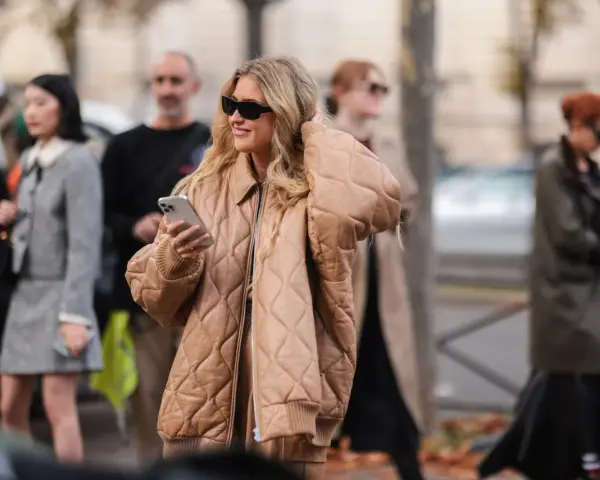 Mujer sonriente con gafas de sol y un abrigo beige claro acolchado, sosteniendo un teléfono.