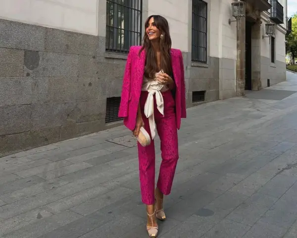 Mujer sonriente luce un espectacular outfit rosa de traje pantalón y blazer, con un cinturón blanco y bolso a juego.