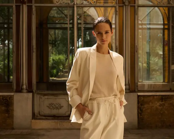 Mujer con blazer beige y pantalones de vestir, de pie frente a una estructura con ventanas.