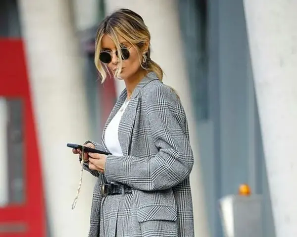 Una mujer con gafas de sol y un traje de cuadros grises, con el pelo recogido, parece una profesora secundaria revisando su móvil.
