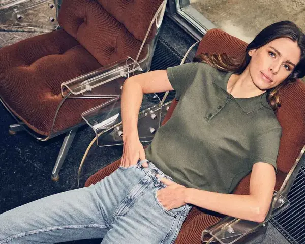 Mujer con vaqueros y polo verde, sentada en una silla de terciopelo, encarnando la moda atemporal.