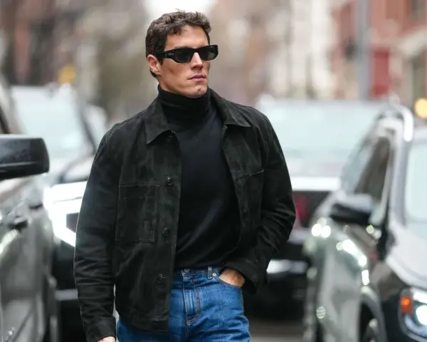 Hombre con gafas de sol y chaqueta de gamuza negra sobre jersey de cuello alto, demostrando como tener estilo.