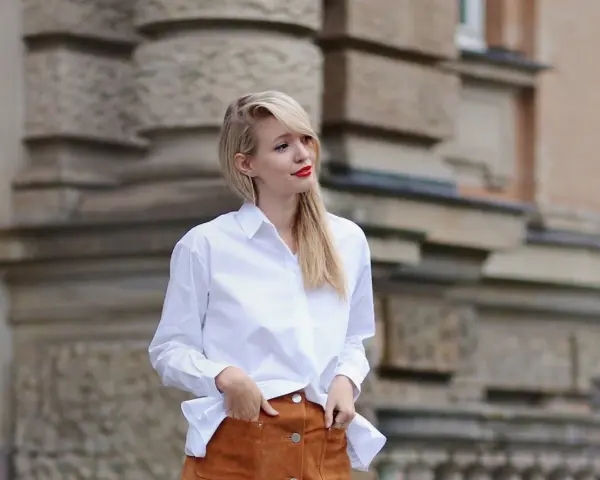 Mujer con falda de gamuza y camisa blanca, posando con las manos en los bolsillos.