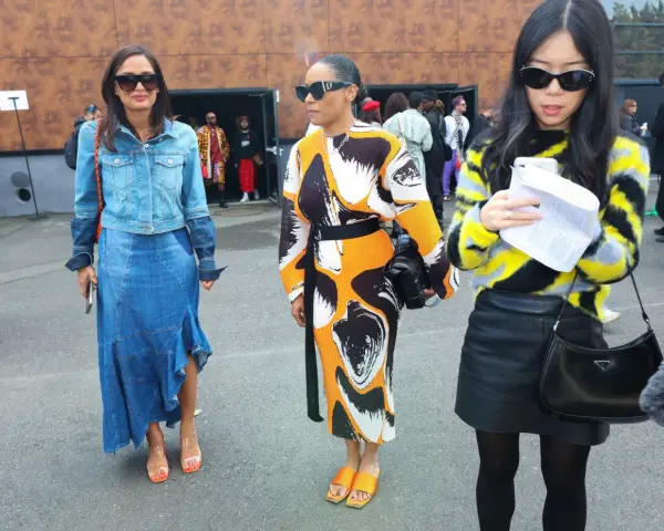 Tres mujeres posan con atuendos de moda. La del centro lleva un vestido estampado y sandalias naranjas, un ejemplo de que son mules.