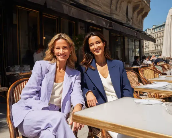 Dos mujeres sonríen sentadas en un café. Una lleva un traje lila, la otra uno azul marino. Ideas para como vestirse para una comunión.
