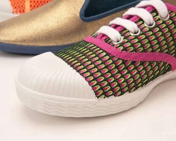 Zapatilla deportiva con malla verde y rosa, puntera blanca y suela vulcanizada. Al fondo, un zapato dorado y otro naranja.