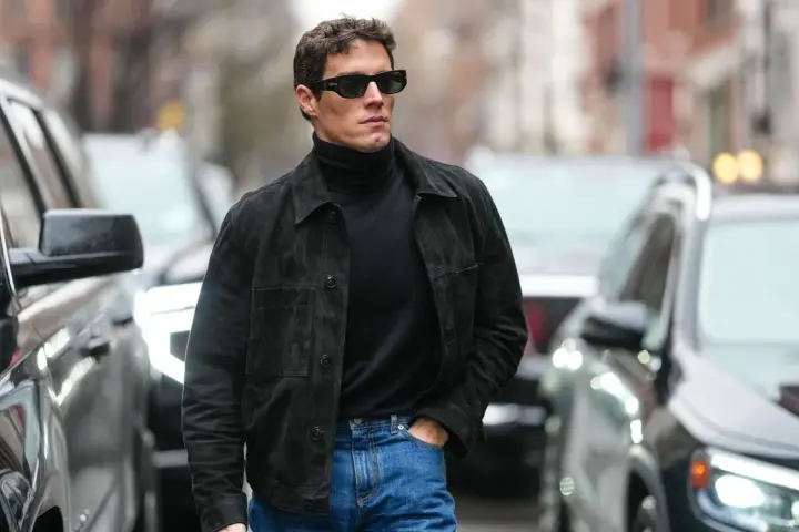 Hombre con gafas de sol y chaqueta de gamuza negra sobre jersey de cuello alto, demostrando como tener estilo.
