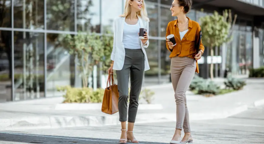 Dos mujeres con looks para ir a trabajar, tomando café y charlando, caminan frente a un edificio moderno.
