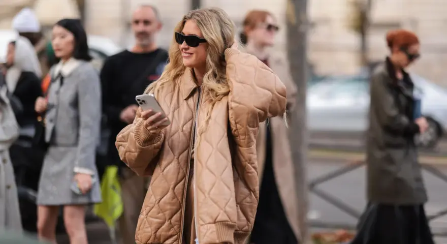 Mujer sonriente con gafas de sol y un abrigo beige claro acolchado, sosteniendo un teléfono.