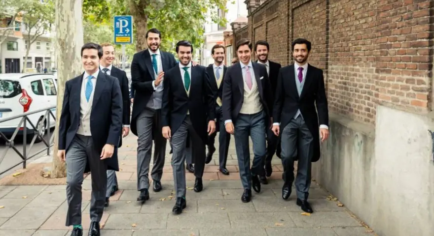 Un grupo de hombres camina por la acera, todos vestidos con trajes elegantes, cumpliendo el **código vestimenta** de un evento formal.