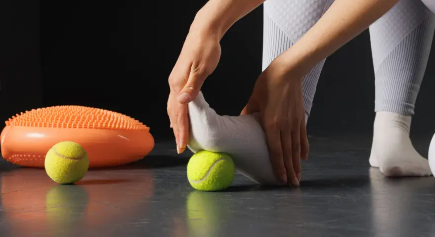 Manos masajeando pie con pelota de tenis, parte de ejercicios fascitis plantar. A su lado, un disco de equilibrio naranja.