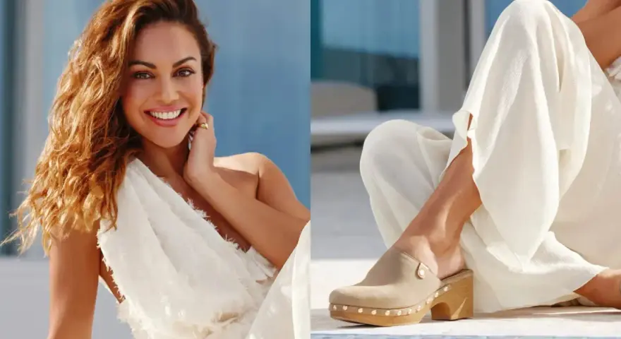 Mujer sonriente con vestido blanco y un pie calzado con zuecos de ante beige, adornados con tachuelas.