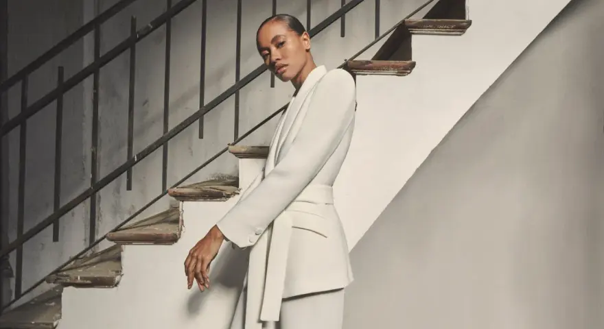 Mujer posando con un elegante outfit blazer blanca, pantalón a juego y cinturón, junto a una escalera antigua.