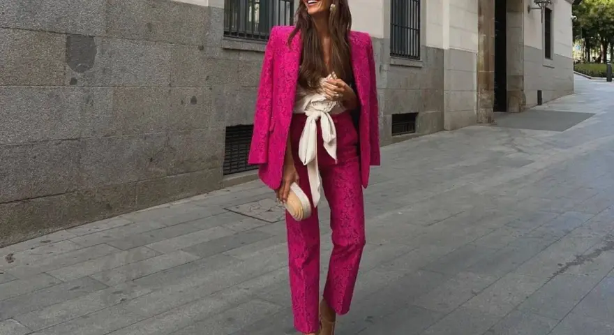 Mujer sonriente luce un espectacular outfit rosa de traje pantalón y blazer, con un cinturón blanco y bolso a juego.