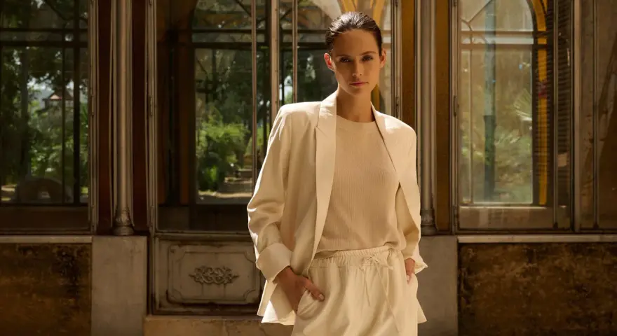 Mujer con blazer beige y pantalones de vestir, de pie frente a una estructura con ventanas.