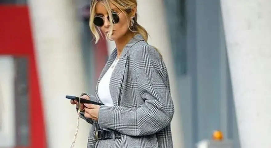 Una mujer con gafas de sol y un traje de cuadros grises, con el pelo recogido, parece una profesora secundaria revisando su móvil.