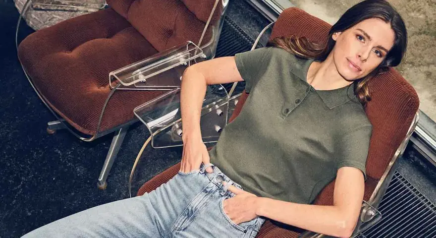 Mujer con vaqueros y polo verde, sentada en una silla de terciopelo, encarnando la moda atemporal.