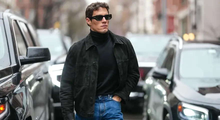 Hombre con gafas de sol y chaqueta de gamuza negra sobre jersey de cuello alto, demostrando como tener estilo.