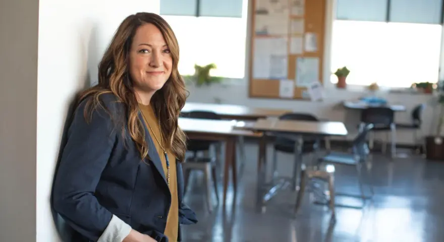Profesora joven sonriente, apoyada en una pared blanca, con un blazer azul marino y un collar llamativo. El aula al fondo está lista para la clase.