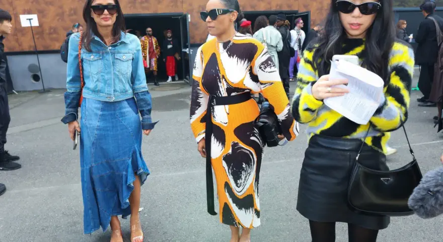 Tres mujeres posan con atuendos de moda. La del centro lleva un vestido estampado y sandalias naranjas, un ejemplo de que son mules.