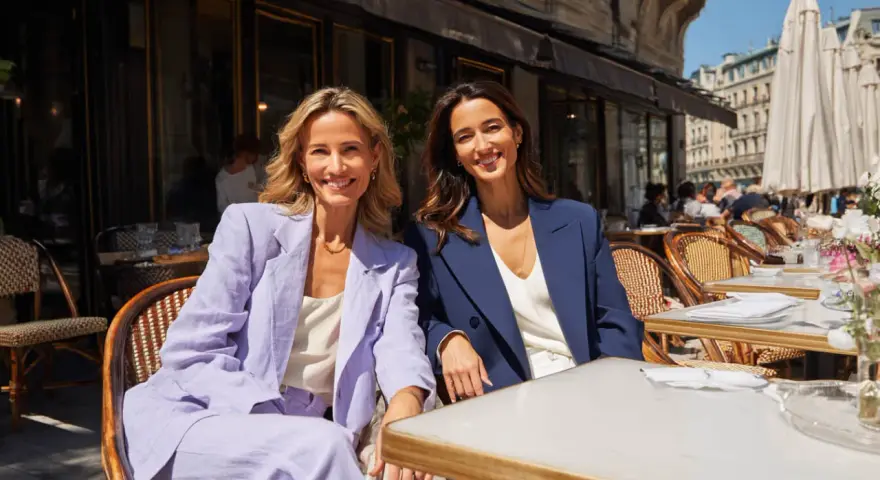 Dos mujeres sonríen sentadas en un café. Una lleva un traje lila, la otra uno azul marino. Ideas para como vestirse para una comunión.