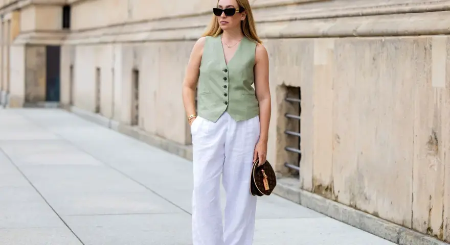 Mujer con un outfit de chaleco verde y outfit pantalon lino blanco, gafas de sol y bolso de mano.