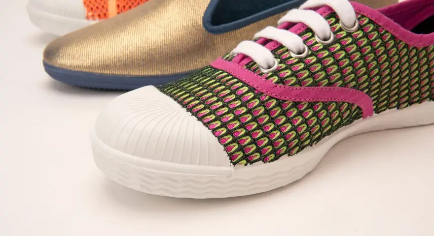 Zapatilla deportiva con malla verde y rosa, puntera blanca y suela vulcanizada. Al fondo, un zapato dorado y otro naranja.
