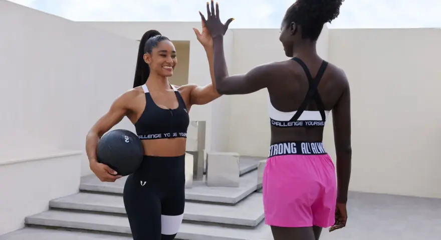Dos mujeres con estilo deportivo se dan la mano en alto. Una sostiene una pesa, la otra lleva shorts rosas.