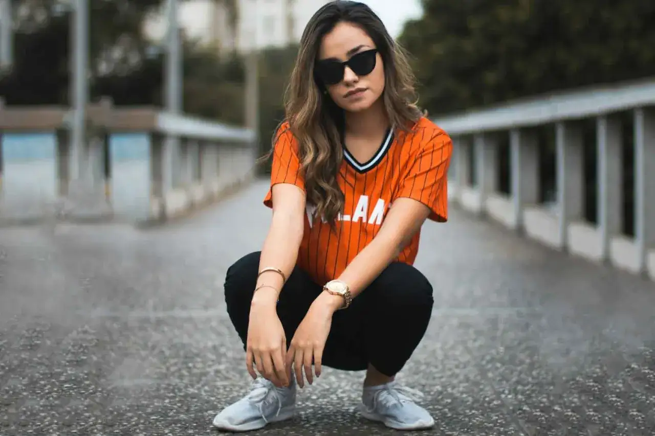 Chica con estilo sporty, gafas de sol oscuras y camiseta naranja a rayas, agachada en un puente.